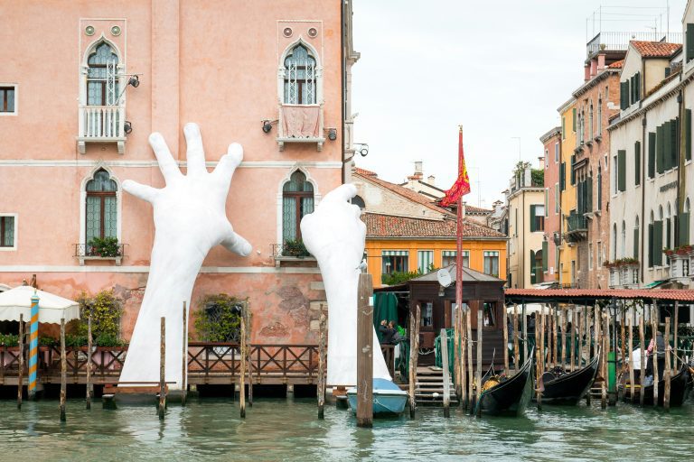 Giant white hand sculpture emerging from canal in Venice near colorful buildings