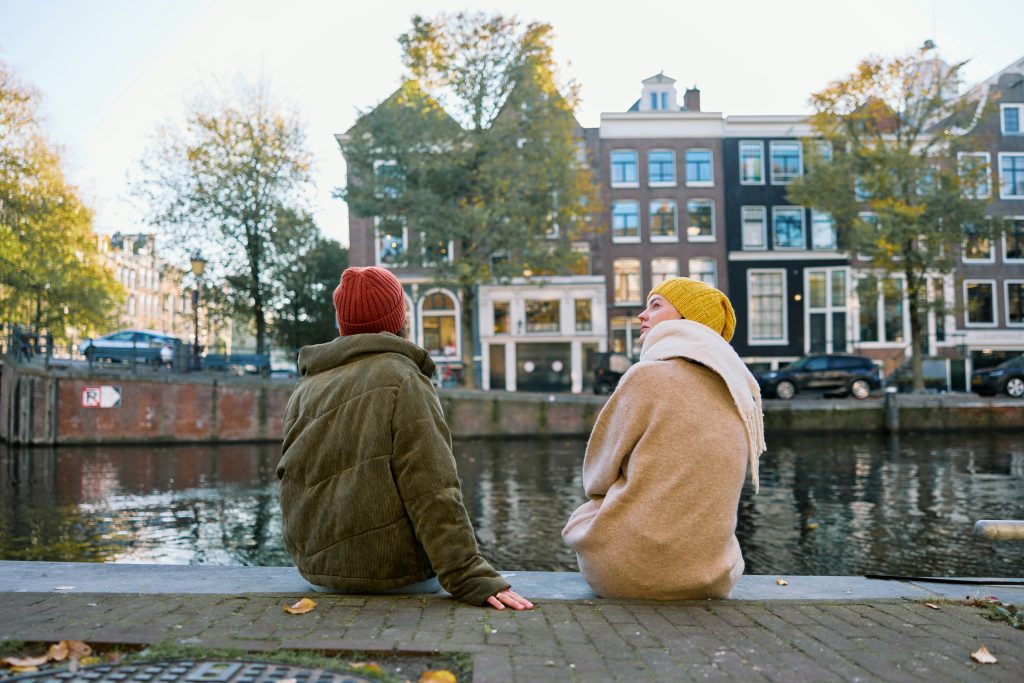 Feiertag in den Niederlanden: zwei Personen sitzen an einer Amsterdamer Gracht mit historischen Häusern im Hintergrund